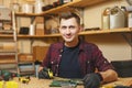 Handsome smiling young man working in carpentry workshop at wooden table place with piece of wood Royalty Free Stock Photo