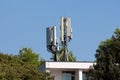 Multiple cell phone antenna transmitters on top of business building surrounded with dense trees with clear blue sky in background