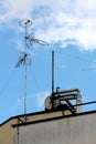 Multiple radio and TV antennas mounted on strong tall metal pole on top of apartment building surrounded with pipes and cloudy