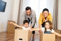 Multiracial family of four have fun in new house, parents riding kids in cardboard boxes in empty living room, happy Royalty Free Stock Photo