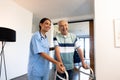 Multiracial female physiotherapist assisting cheerful senior man in walking with walker at home Royalty Free Stock Photo