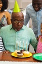 Multiracial senior man wearing hat blowing birthday candles while celebrating with family at home Royalty Free Stock Photo