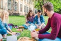 multiracial smiling students with digital devices sitting on green grass