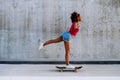 Multiracial teenage girl riding a skateboard in front of concrete wall, standing in one leg, balancing. Side view.