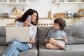 Busy businesswoman mother work from home talk to kid with laptop on knees calling on phone on couch