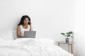 Multitasking. Happy black woman talking on smartphone and using laptop computer, sitting on bed at home, empty space Royalty Free Stock Photo