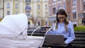 Multitasking mother swinging baby carriage and working laptop sitting on bench Royalty Free Stock Photo