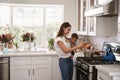 Multitasking mum holding her young baby while she makes food at the hob in her kitchen, side view Royalty Free Stock Photo