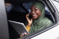 Multitasking. Smiling black muslim woman using digital tablet and smartphone in car Royalty Free Stock Photo