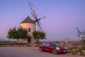 Murcia, Spain - October 26, 2019: A red Tesla Model 3 is parked in front of a windmill surrounded by trees. Its headlights are on