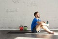 Muscular athlete sitting in sunny sport club, relaxed after workout, bottle of water in hands. Space for text layout on brick wall