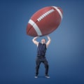 A muscular athletic man has trouble to hold a giant American football ball over himself.