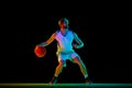 Muscular, athletic young man, basketball player in motion with ball, practicing against black background in neon light