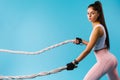 Muscular girl throws the waves of the battle rope to the side and looks at the camera on a blue background. Sports power