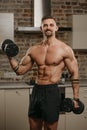 A muscular man with beard and tattoos is training biceps with dumbbells at home