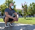 Muscular man crouched down in a public park wearing a face mask, with a lot of space on the right side Royalty Free Stock Photo