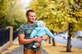 A muscular man holds the baby in warm jumpsuit in the fall on a walk. yellow leaves