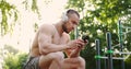 Muscular man sitting on sports ground in city park and checking his phone while listerning to music