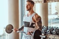 Muscular tattooed man is doing his exercises with barbell in gym near window