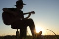 Music concept of black silhouette of man with western hat cowboy and instrument at sunset or sunrise with skyline and grass.