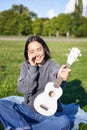 Music and instruments. Smiling asian girl shows her white ukulele, sits in park and plays small guitar Royalty Free Stock Photo