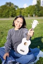 Music and instruments. Smiling asian girl shows her white ukulele, sits in park and plays small guitar Royalty Free Stock Photo