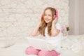 Music and technology. Portrait of young girl in pajamas with white laptop and wireless pink headphones on background