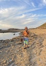 A musician saxophonist with a naked torso in bright multi-colored wide pants and headphones walks along the seashore with a