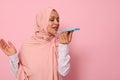 Muslim Arab beautiful gorgeous woman with covered head in pink hijab recording a voice message on her smartphone. Isolated