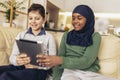 Muslim girl and her caucasina friend sitting on sofa at home using digital tablet