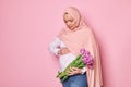 Muslim pregnant woman in pink hijab, touches her tummy, feels first baby kicks, holds beautiful tulips, isolated on pink