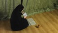 A Muslim woman in a black khimar in profile at home reads the Holy Quran on a rug