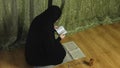 A Muslim woman in a black khimar in profile at home reads the Koran on a rug