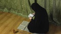 A Muslim woman in a black khimar in profile at home reads the Koran on a rug