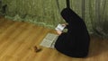 A Muslim woman in a black khimar in profile at home reads the Koran on a rug