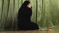 A Muslim woman in a black khimar reads prayer on her knees on a rug at home