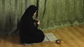 A Muslim woman in a black khimar reads prayer on her knees on a rug at home