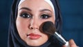 Muslim woman doing makeup on her face with brush.