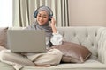 Muslim woman wearing hijab and headphones to listen the music and using laptop in living room Royalty Free Stock Photo