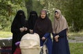 Muslim women wearing hijabs standing on a park background, one holding a baby buggy