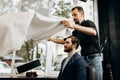 Mustachioed barber dressed in a black shirt puts a cape on the stylish man sitting in the armchair in a barbershop