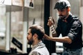 Mustachioed barber dressed in a black shirt and a red bow tie is doing the hair styling to the stylish man sitting in