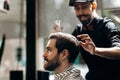 Mustachioed barber dressed in a black shirt and a red bow tie is doing the hair styling to the stylish man sitting in