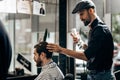 Mustachioed barber dressed in a black shirt, red bow tie and flat cap is doing the hair styling to the man sitting in