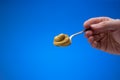 Mustard paste on a small silver spoon held in hand by man. Close up studio shot, isolated on blue background Royalty Free Stock Photo