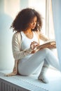 Calm relaxed african american young woman reading book while sitting on windowsill at home