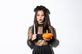 Myserious asian wicked witch in gothic dress, looking at lit candle, holding pumpkin, standing over white background