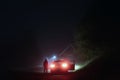 A mysterious man standing next to a car on a spooky empty road on a foggy night. Silhouetted by street lights. Royalty Free Stock Photo