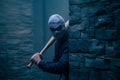 Mysterious figure with baseball bat hides behind stone wall in eerie atmosphere.
