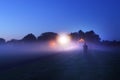 A mysterious, ghostly, lone man standing on a path in a field. Looking at street lights. Just before sunrise on a misty early